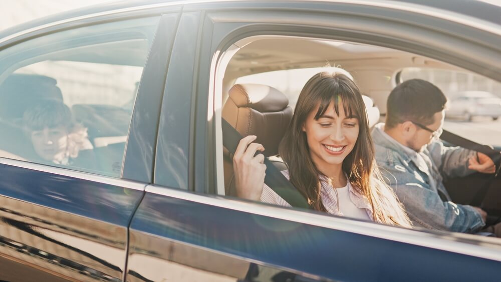 A teen buckles her seatbelt with a warm smile while riding in the car with her family, capturing a safe and reassuring moment supported by teen driver insurance.