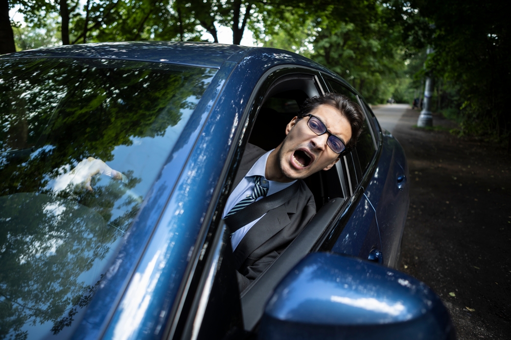 Hombre molesto asomado por la ventana del auto en la calle, mostrando frustración porque otro auto obstruye mi entrada.