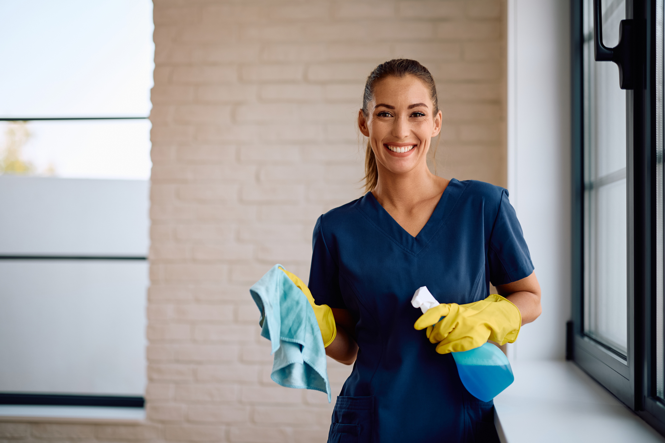 Mujer sonriente con uniforme azul y guantes amarillos sosteniendo un paño de limpieza y una botella con atomizador azul, junto a una ventana en un espacio interior luminoso.