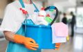 Persona con delantal azul y guantes naranjas sosteniendo un balde azul lleno de productos de limpieza, incluyendo botellas con atomizador, detergentes, esponjas y paños de microfibra, en un entorno interior desenfocado.