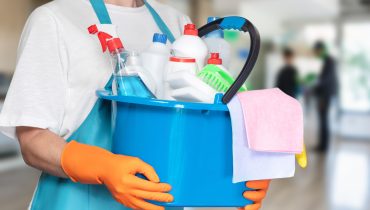 Persona con delantal azul y guantes naranjas sosteniendo un balde azul lleno de productos de limpieza, incluyendo botellas con atomizador, detergentes, esponjas y paños de microfibra, en un entorno interior desenfocado.