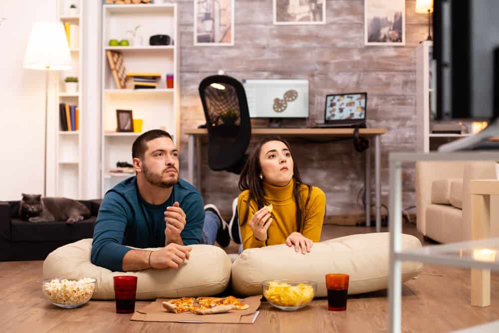 Pareja comiendo pizza y snacks mientras mira la televisión en casa, representando la comida típica en NASCAR.