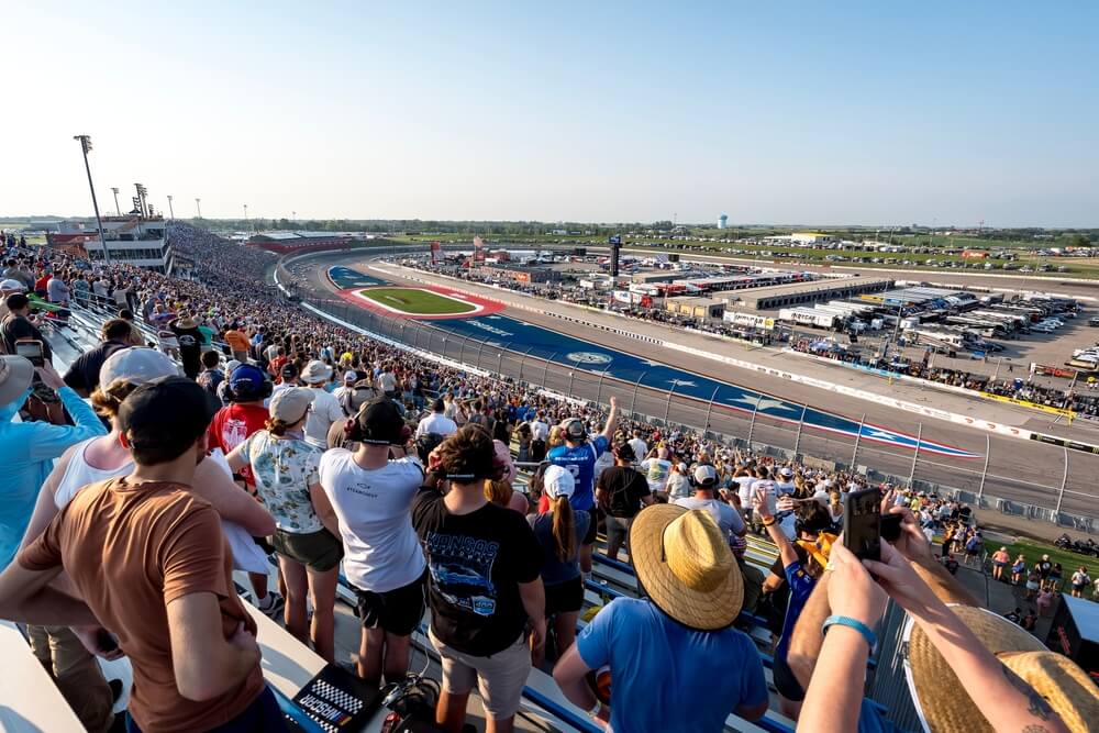 Aficionados disfrutando de una carrera de NASCAR desde las gradas de un gran autódromo, mostrando la emoción y la experiencia familiar del evento.