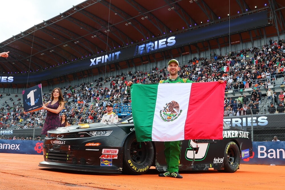 Daniel Suárez posa con la bandera de México junto a su auto de carreras, simbolizando el impacto latino en NASCAR y el orgullo de la comunidad hispana en este deporte.