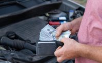 Persona sostiene dinero frente al motor de un automóvil, ilustrando el reto de cubrir reparaciones con ahorros propios en lugar de contar con un seguro de auto.