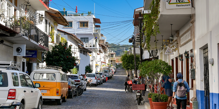 Calle empedrada en pueblo mexicano con autos estacionados y personas caminando, ideal para seguro de auto para viajar a México.