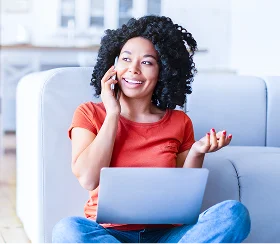 Una mujer sonriente está sentada en un sofá usando una laptop mientras habla por teléfono, ilustrando comparaciones inteligentes lado a lado y ahorro de tiempo.