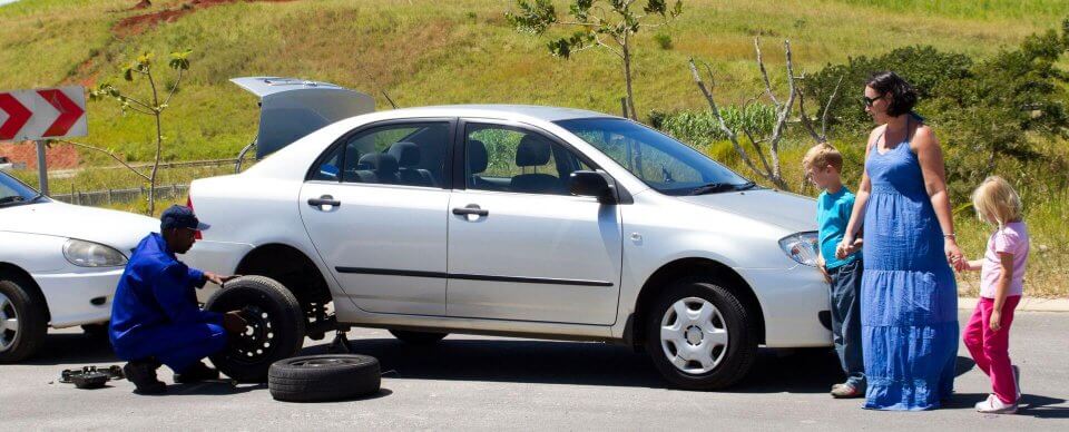 A multi-ethnic family that had a flat waits as a road-assistance worker changes the damaged tire.