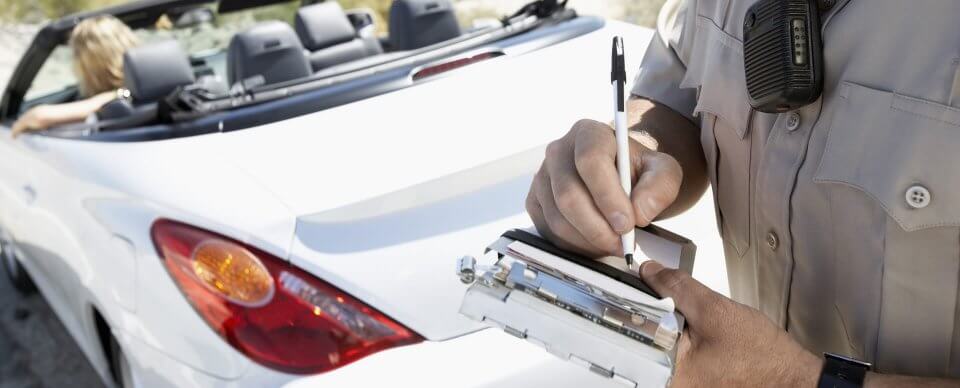 A traffic officer writing a ticket for a young person driving in a convertible car to illustrate how an out-of-state speeding ticket does matter