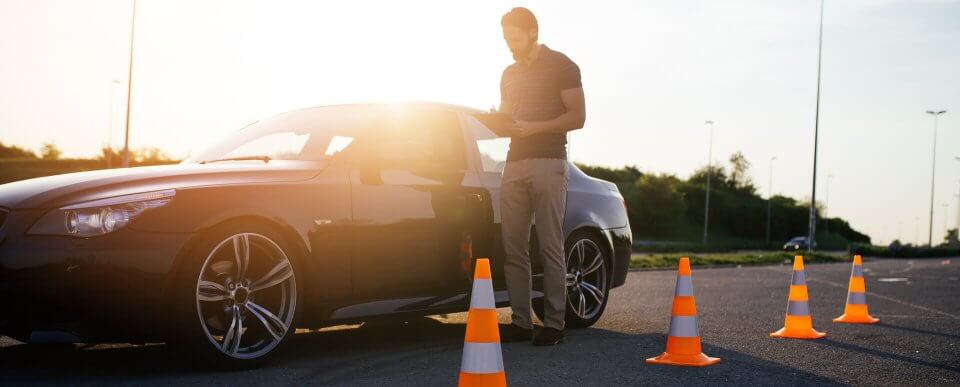 A driving instructor standing next to a coupe car in a traffic course to illustrate reasons to take a defensive-driving course.