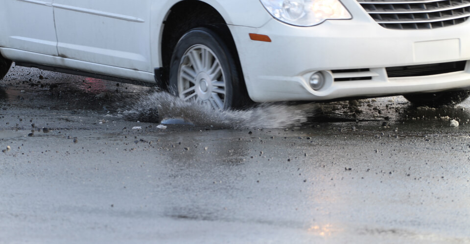 A car hitting a pothole on a rainy road to illustrate how to avoid potholes and prevent costly repairs.