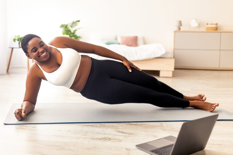 Online sports for weight loss. Overweight black woman doing exercises in front of laptop, standing in side plank at home. Plus size young lady following video tutorial, training indoors.