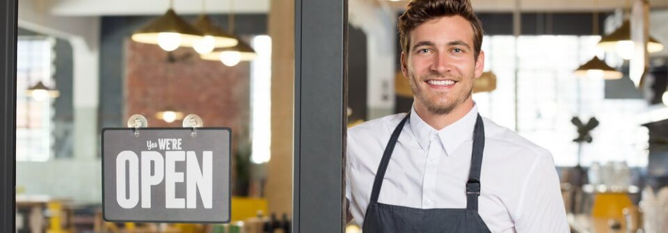 Portrait of smiling business owner standing at his restaurant gate with open signboard to portray 3 benefits of getting business insurance