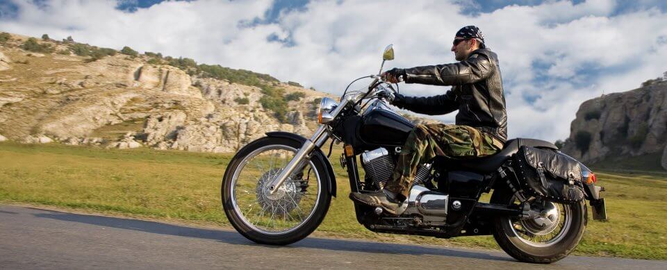 A middle-age Caucasian man riding his chopper motorcycle down a lonely road thanks to the appropriate motorcycle insurance.