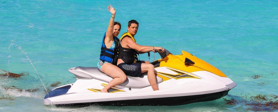 A middle-age couple riding a jetski and waving at the camera to portray reasons to buy watercraft insurance.