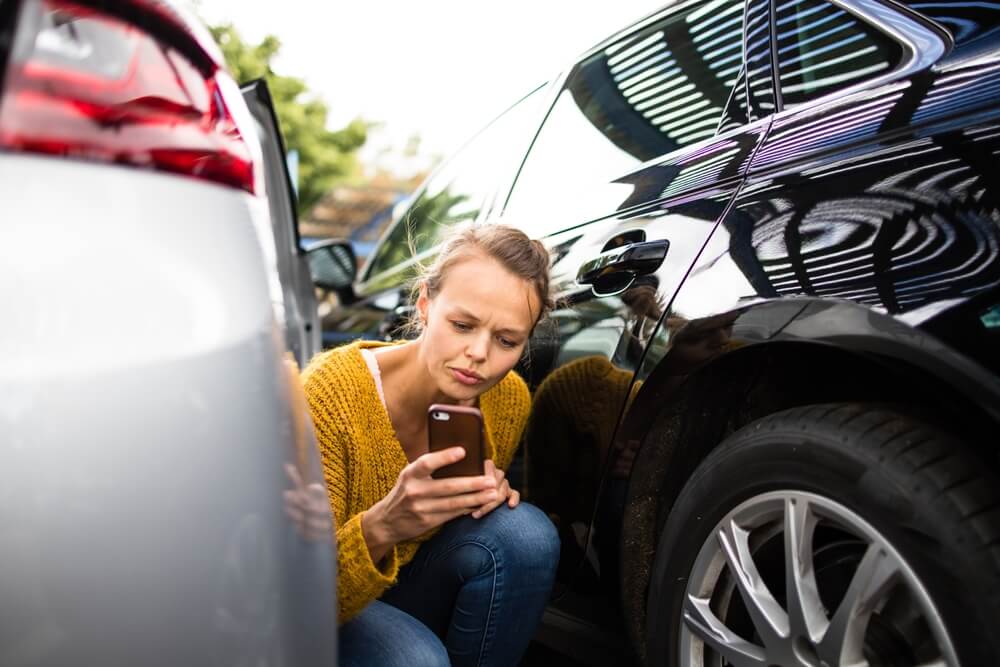 Worried woman crouches between two cars after a minor accident, checking her phone for steps on how to file a car insurance claim.