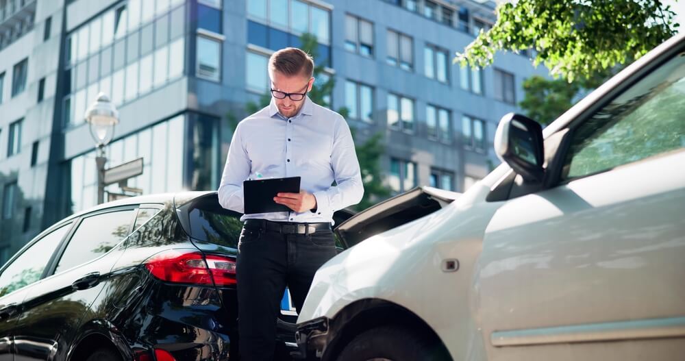 Insurance adjuster in a button-up shirt and glasses inspects the damage between two cars and takes notes on a clipboard, showing how to file a car insurance claim.