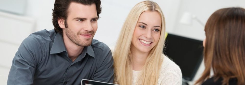 A young caucasian couple smiling at their insurance broker to illustrate what is a brokerage fee and why it should be paid.