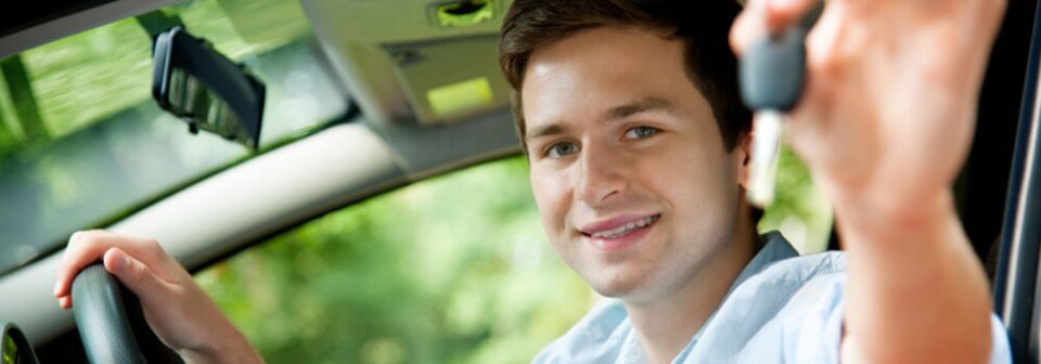 A young Caucasian driver holds his car keys out of his car's window to illustrate how to help your teen driver stay safe.