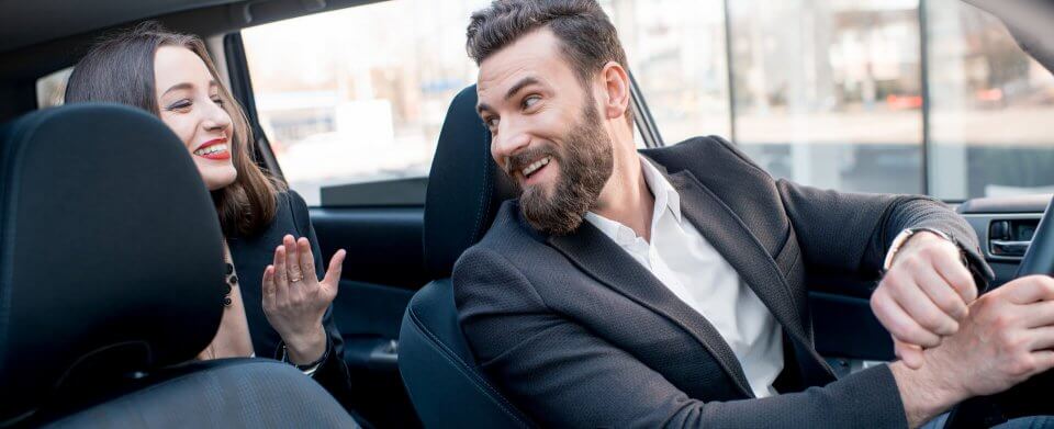 A young, Caucasian ridesharing driver who has commercial vehicle insurance smiles at a passenger in the backseat.