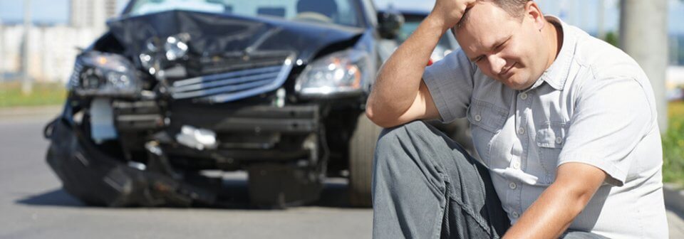 A middle-age Caucasian man sitting in front of his totaled car in regret.