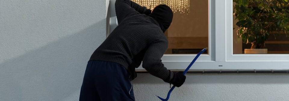 A man in black attire using a crowbar to bust open a window portrays ways to protect your home from break-ins and burglaries.
