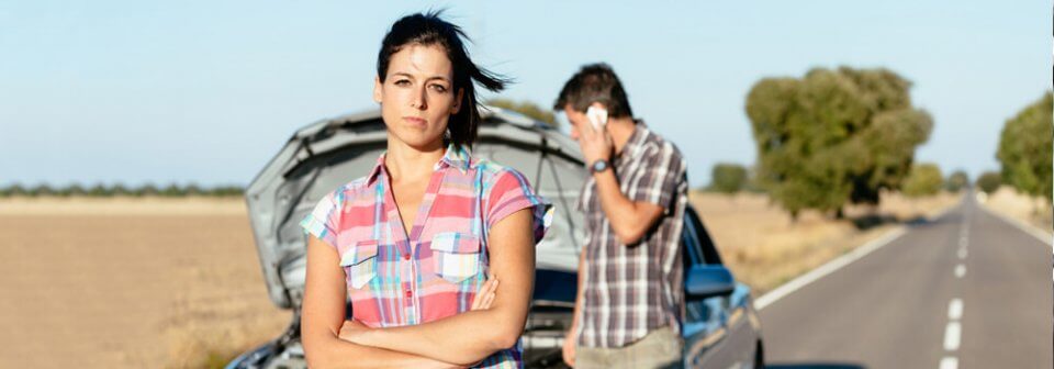 A young, Caucasian woman stares in anger at the horizon as a man in the back makes a phone call because of a ruined car.