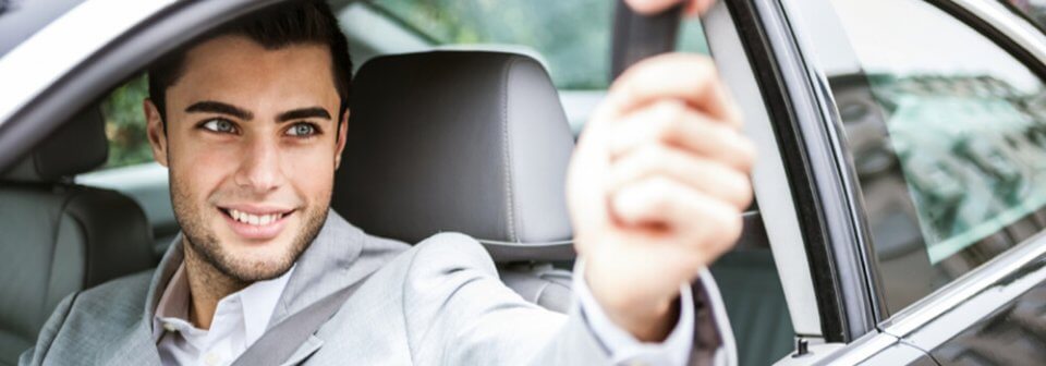 A young Caucasian male sitting on the driver's seat of a car receives the car keys from the rental agent to illustrate whether insurance will cover a car rental