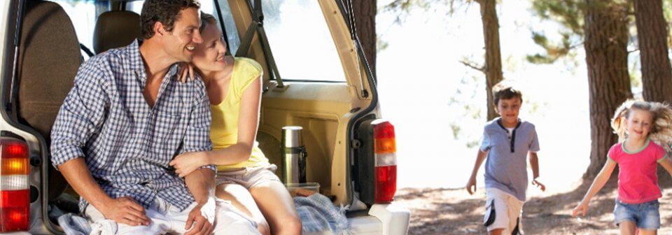 A young couple sitting in the back of their station wagon while its children run and play during a holiday.