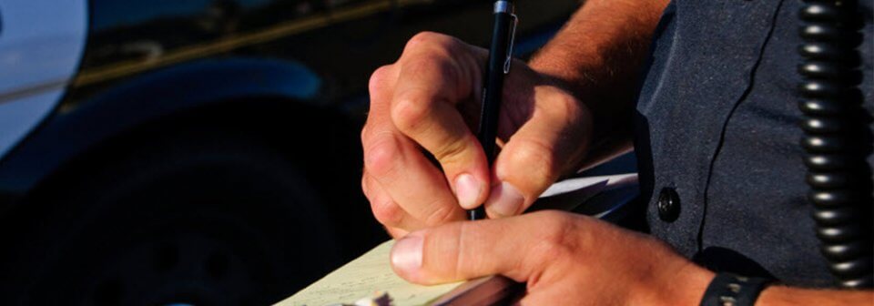 Close-up to the hands of a traffic officer as he writes a ticket for a driver to illustrate how two different points systems affect your auto-insurance rate.