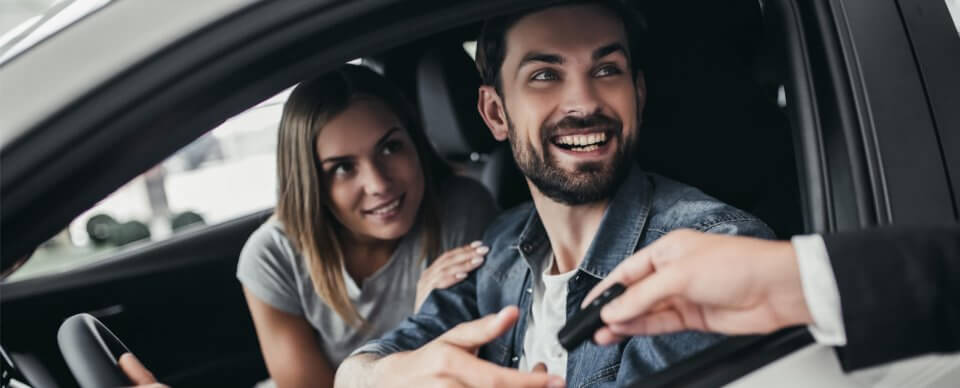 A young couple who just purchased their first car smiles at the car sales agent who hands them the keys to it. They have new car insurance.