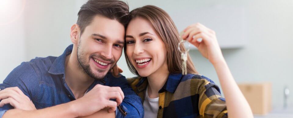 A young Caucasian couple smiling and holding the keys to their new home. The have homeowners' insurance for the first time home-buyer.