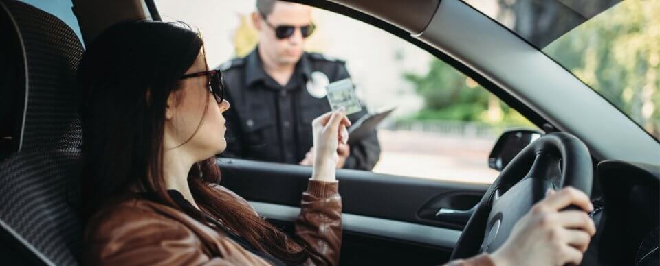 An annoyed Caucasian woman showing her license and SR-22 proof of financial capacity to a traffic officer who pulled her over.