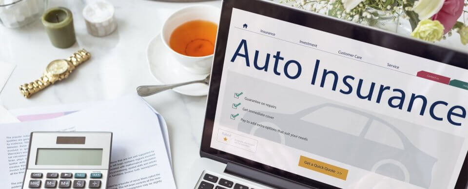 A laptop displaying information on auto insurance placed next to a policy, tea and a calculator to portray an excluded driver on insurance.