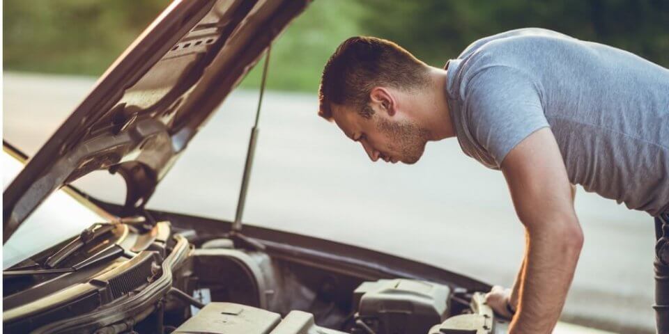 A worried man looking at the engine of a broken down car to illustrate why do you need a roadside assistance plan.