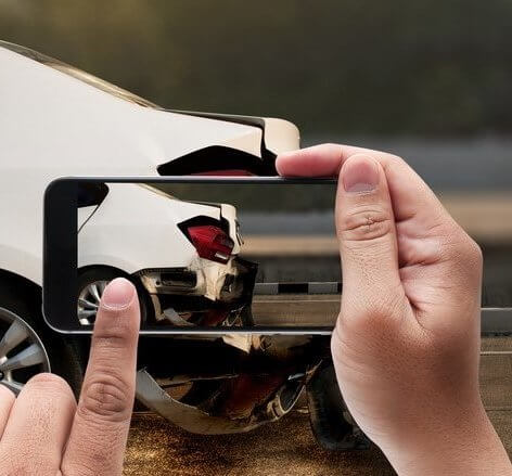 Close up to a pair of hands taking a picture of a damaged bender with a smart-phone to illustrate how to report a hit-and-run incident.