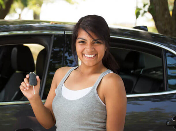A young hispanic teen driver stands in front of her car to illustrate quality insurance for teenage drivers.
