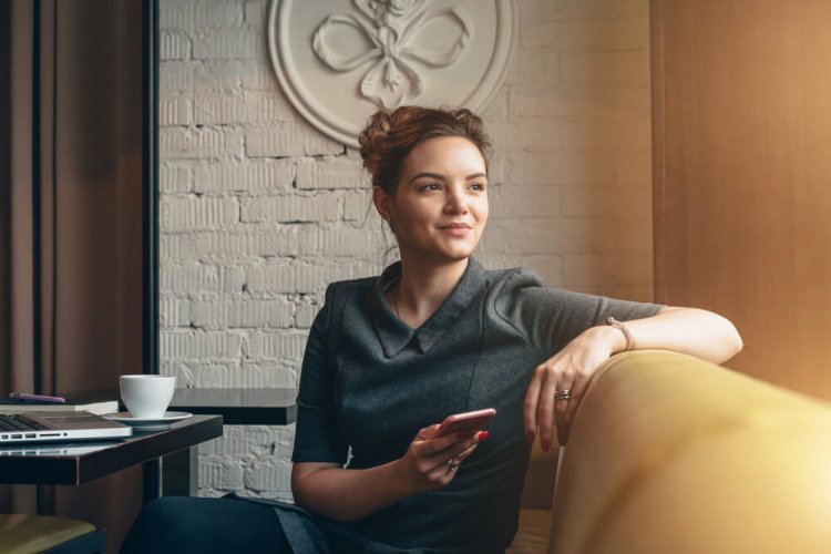 young woman looking out her living room window with a smartphone on her hand