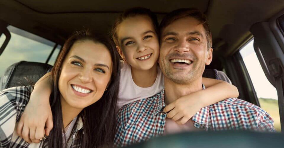 A kid hugging both her parents, smiling in the car.