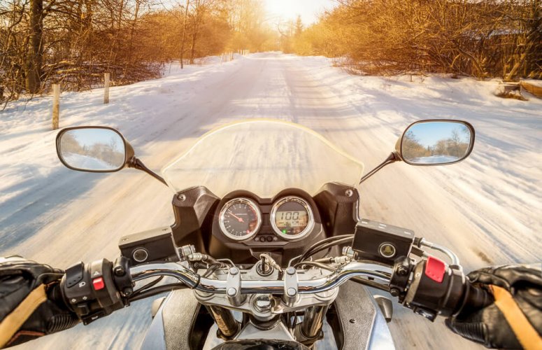 Point of view of a motorcycle rider driving on a quiet, lonely road covered in snow in a forest to illustrate the need for motorcycle insurance year-round.