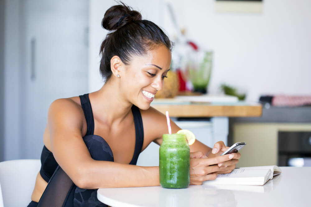 young atlethic woman smiling with green smoothie and looking at cell phone for cheap life insurance