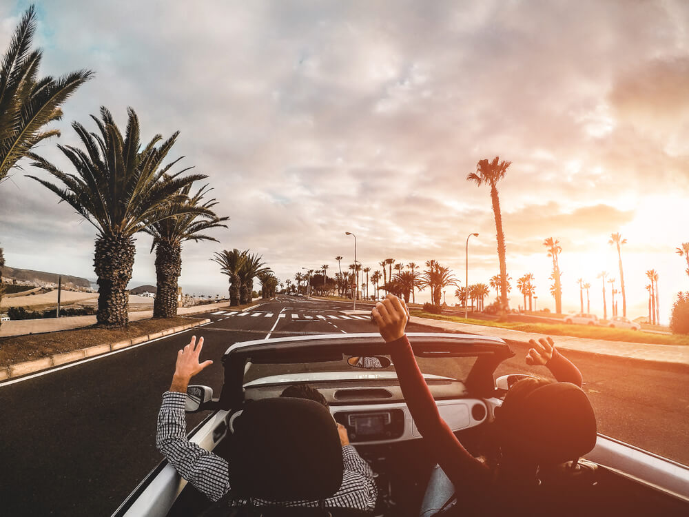 two people driving in car with palm trees in distance and hands in the air