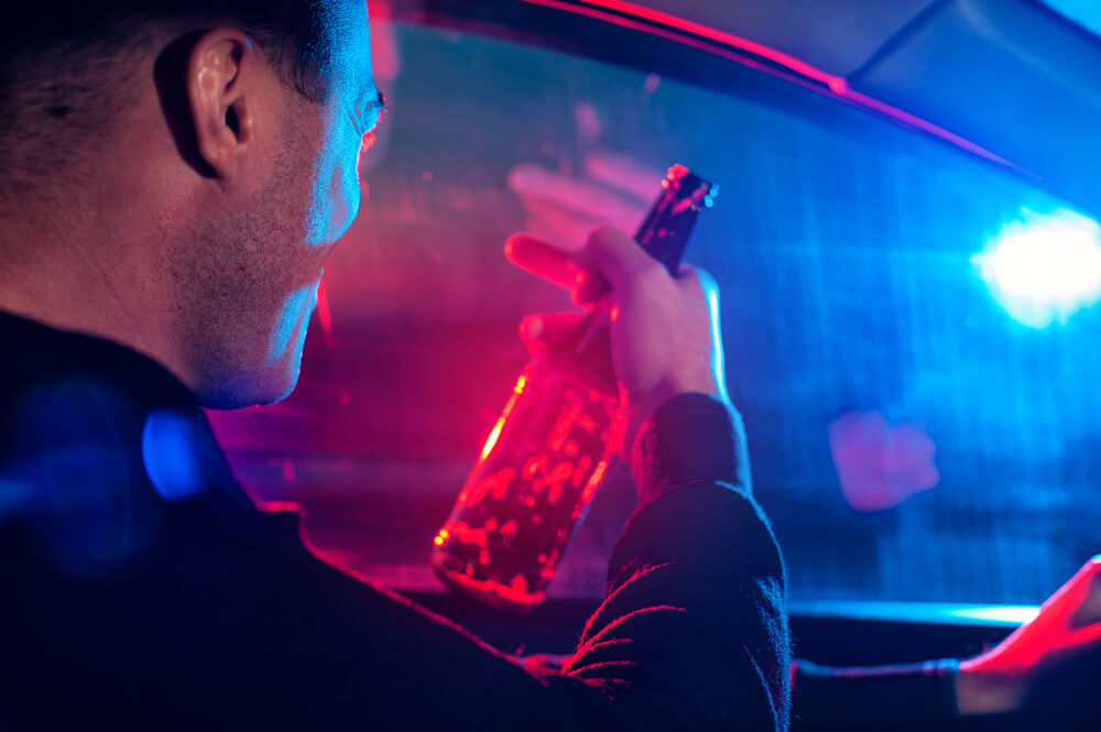 Man in car holding beer bottle behind the wheel with red and blue lights