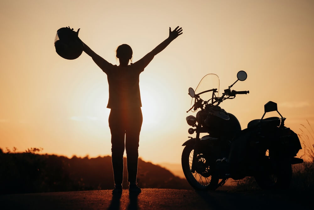 Silhouetted rider raises a helmet beside a parked motorcycle at sunset, highlighting Motorcycle Helmets and Insurance.