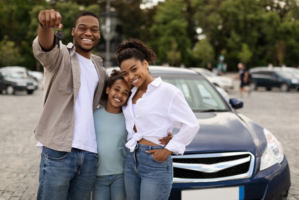 African American man, woman and child with car keys on a car lot in front of a car