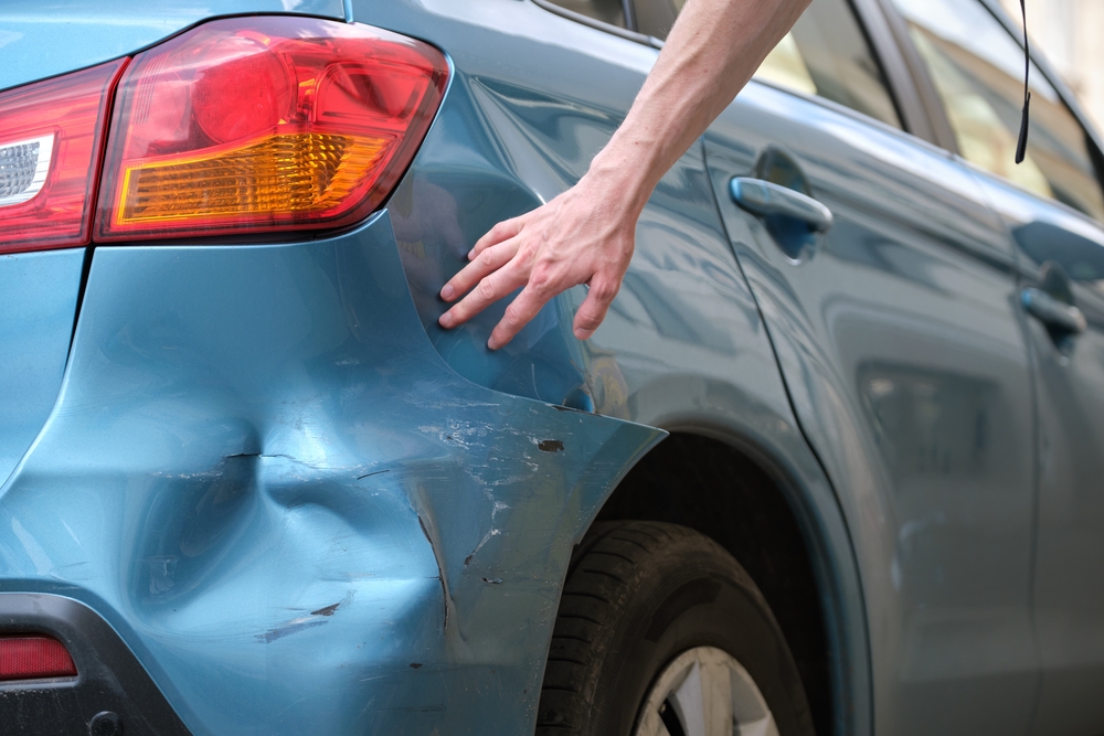 Person inspecting a dent and scratches on the rear bumper of a blue car.