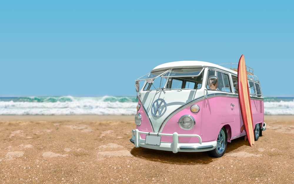 An old-style pink Volkswagen Bus at the beach with a surfboard