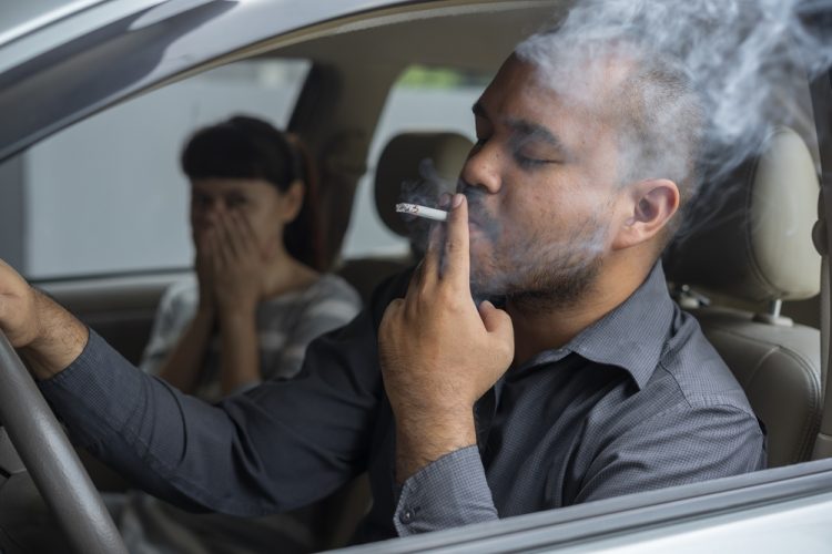 Man smokes in the car while woman in the passenger seat grimaces