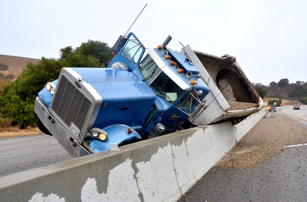 A dump truck on its side on a concrete barrier