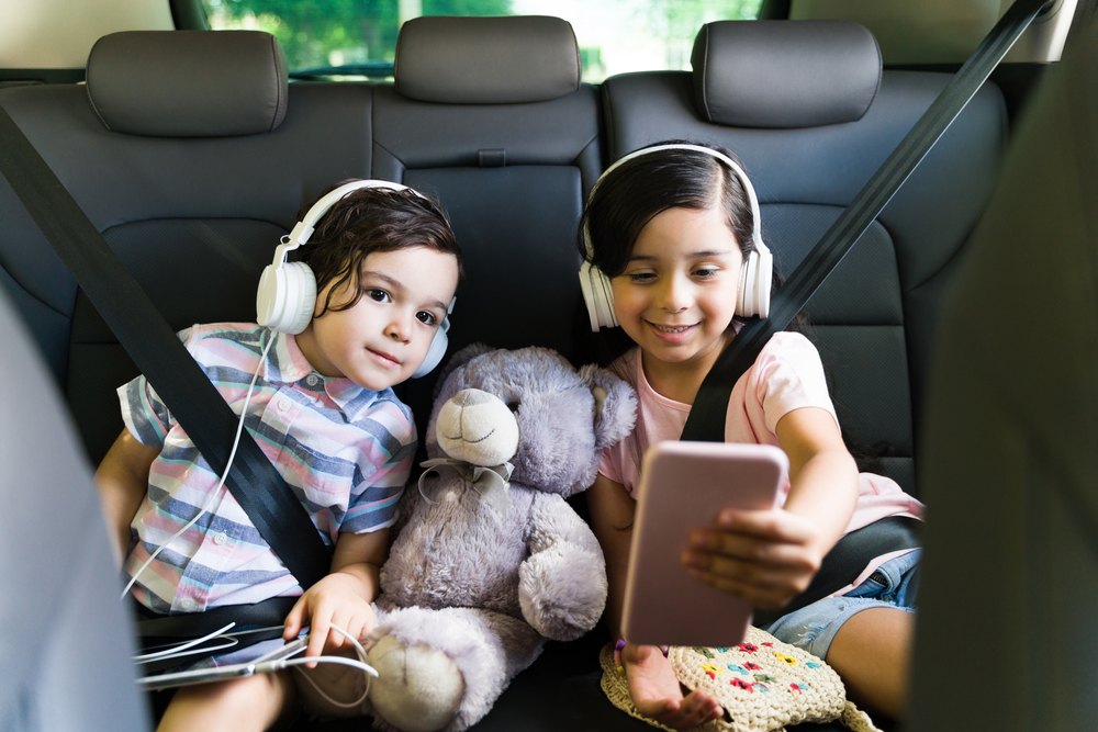 Two adorable children in the back seat of the car with phone, teddy bear and headphones